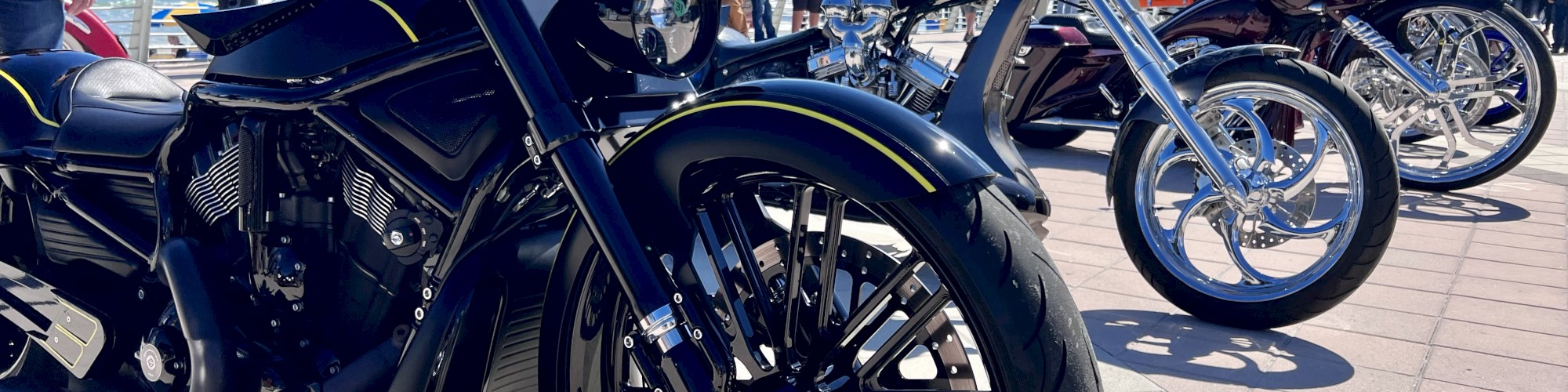 Custom motorcycles lined up outdoors with people in the background under a clear blue sky.