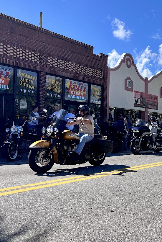 The image shows a street lined with motorcycles and riders near a bar and grill, under a sunny sky with palm trees and passing clouds.