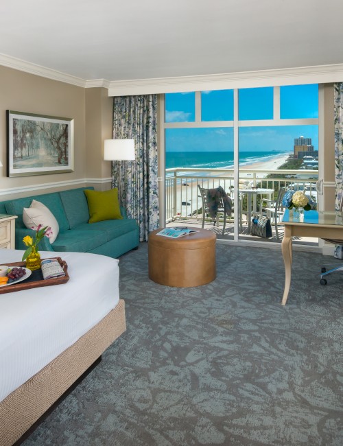 A hotel room with a bed, couch, desk, and a window view of the ocean. A tray with food is on the bed, and there are two lamps.