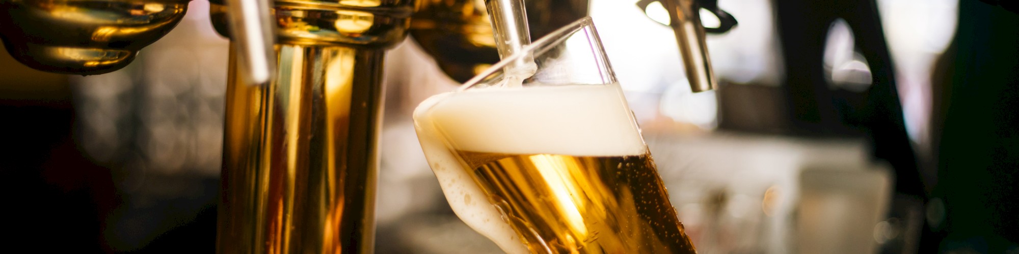 A person pouring beer from a tap into a tilted glass, creating a foamy head. The setting is likely a bar or a pub environment.