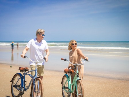 A couple is enjoying a leisurely bike ride along a sunny beach with waves in the background.