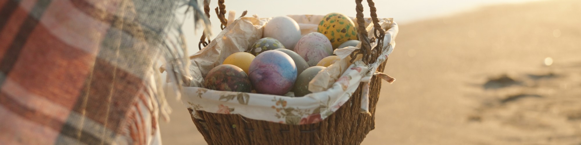 A person is holding a basket filled with colorful Easter eggs on a sandy beach at sunset.