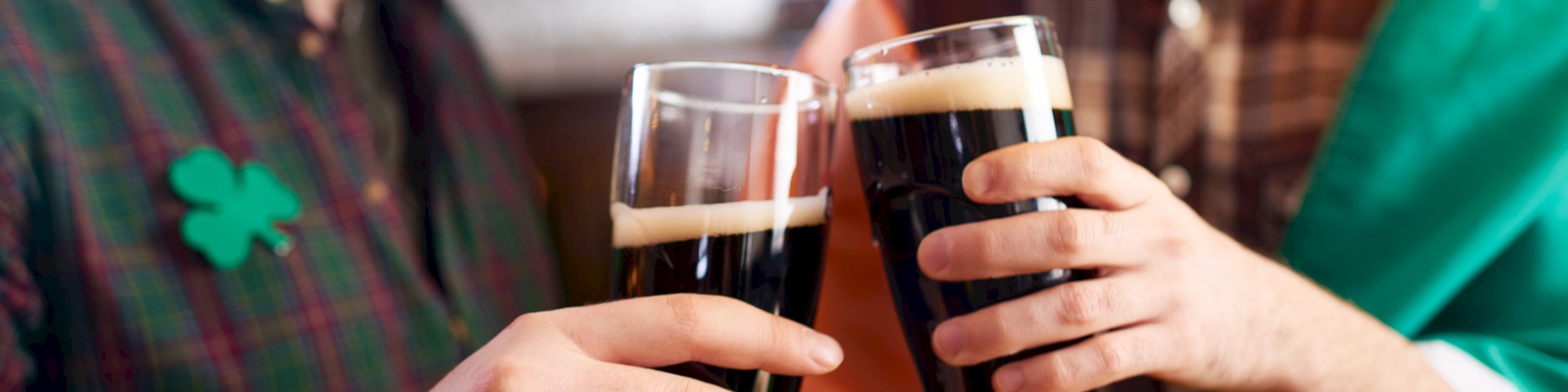 Two people clinking glasses of dark beer, wearing festive attire with an Irish theme, including a shamrock and flag colors.