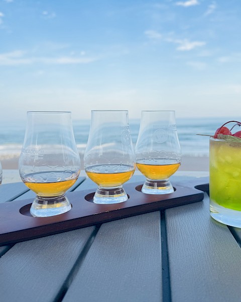 A flight of four whiskey glasses and a cocktail with cherries, set on a table by the beach under a clear blue sky.