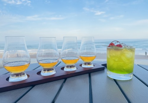 A flight of four whiskey glasses and a cocktail with cherries, set on a table by the beach under a clear blue sky.