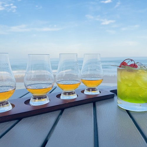 A flight of four whiskey glasses and a cocktail with cherries, set on a table by the beach under a clear blue sky.