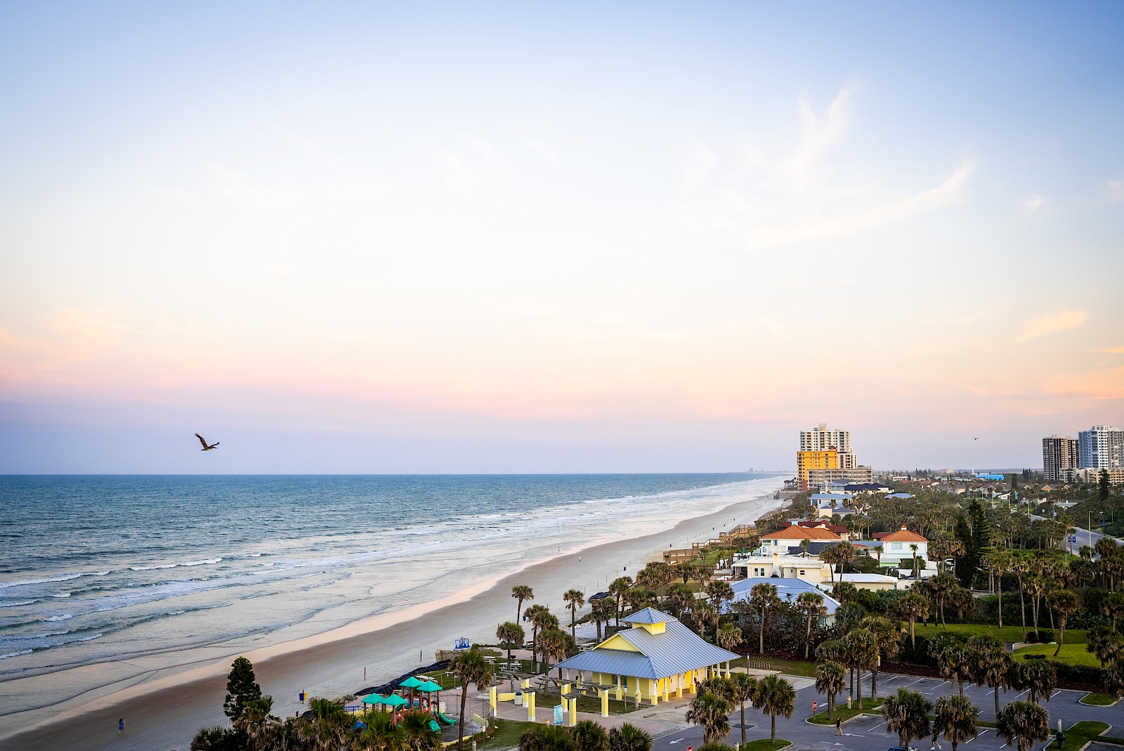 A sunny coastal scene with a beach, ocean waves, and palm-lined hotels along the shore under a clear sky.