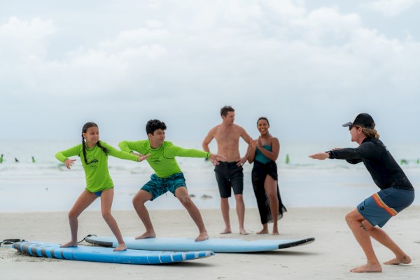 A group on a beach learns to balance on surfboards, two kids in neon rash guards practice with a coach cheering from the side, windy skies.