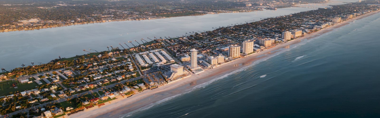 Aerial view of a coastal city with high-rise buildings along the beach, next to a body of water and dense urban area in the distance.