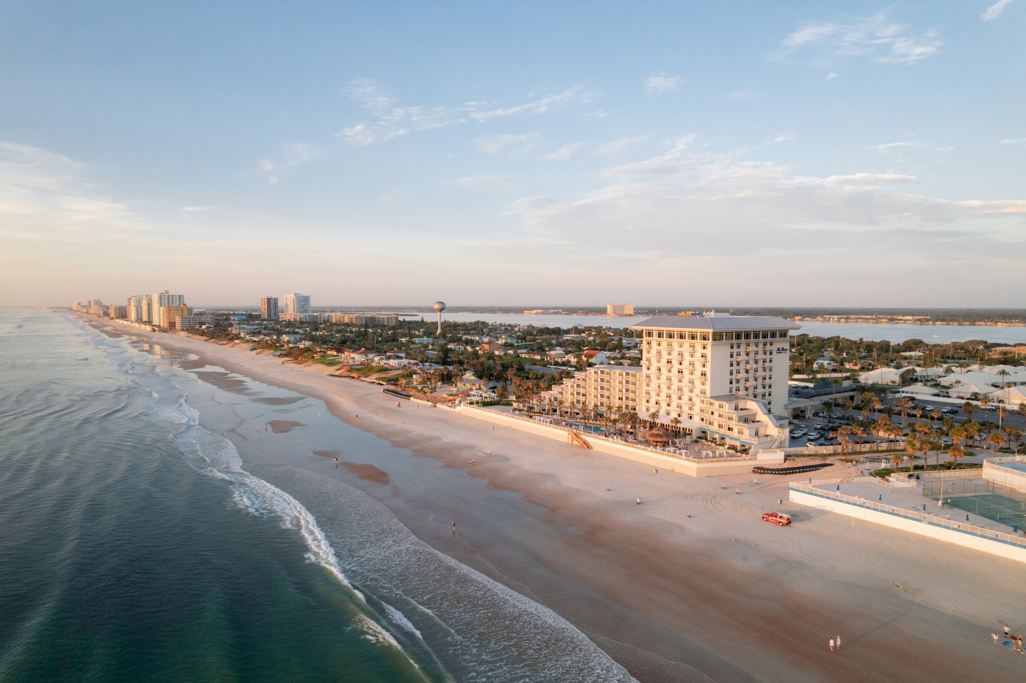 Aerial view of a coastal city with a sandy beach, gentle waves, beachfront buildings, and a clear sky in the background.