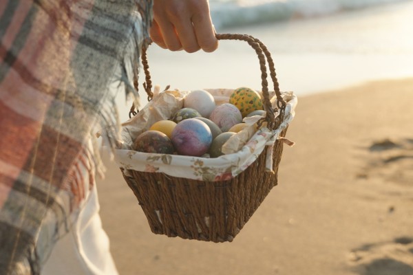 A person is holding a basket filled with colorful, decorated eggs while walking on a beach.