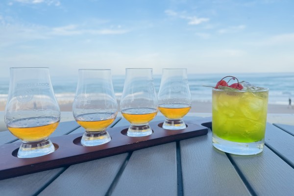 A flight of four whiskey glasses and a green cocktail with cherries on a beachside table overlooking the ocean.