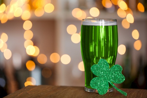 A glass of greened-colored drink with a glittery shamrock decoration, celebrating St. Patrick’s Day with blurry warm lights in the background.