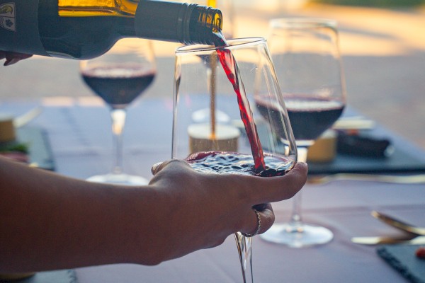 A bottle of red wine being poured into a glass on a table set with utensils and other filled wine glasses overlooking the ocean.