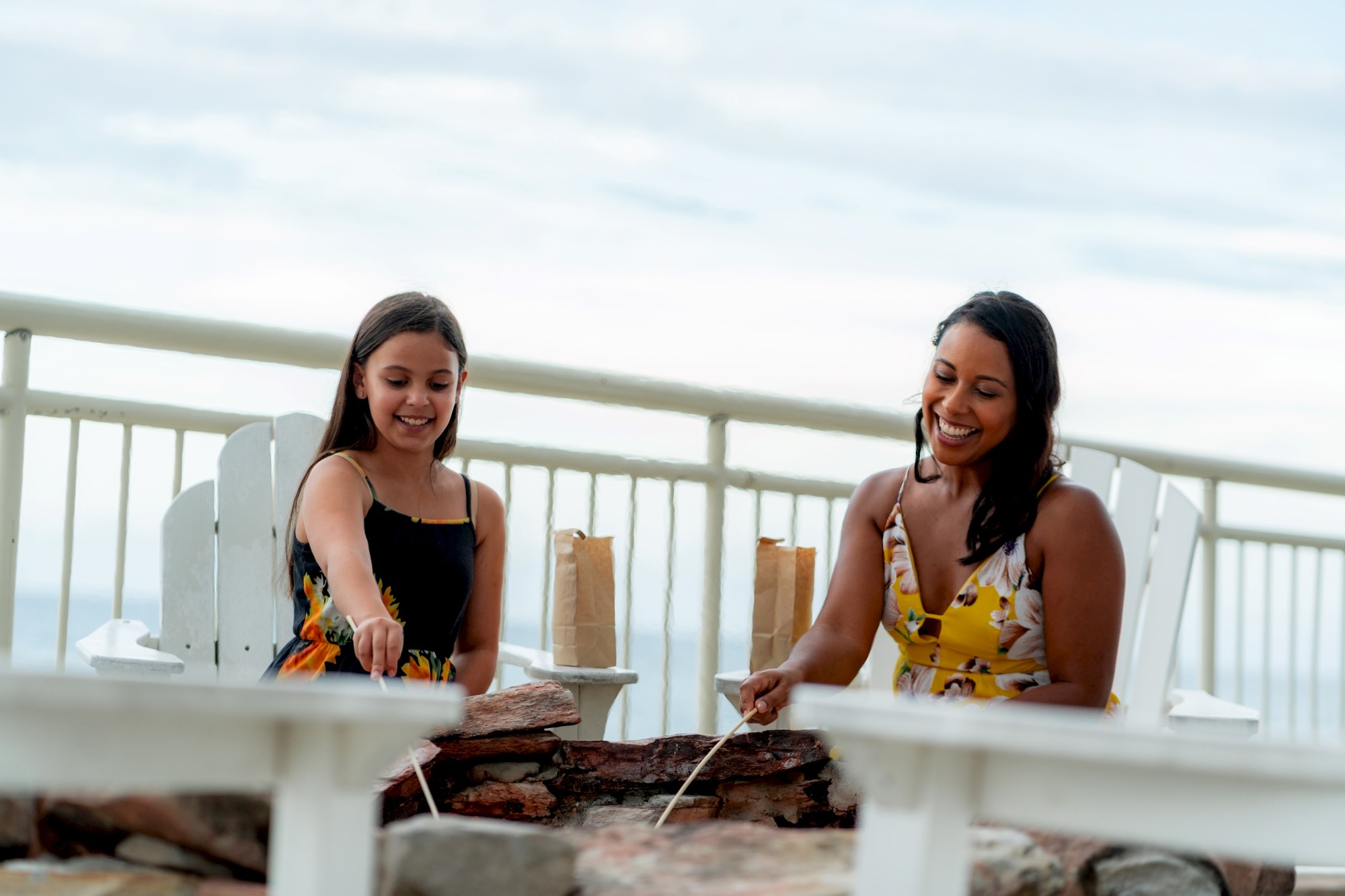 A woman and a girl sit by a fire pit, roasting marshmallows. They are smiling and enjoying a relaxed outdoor setting.