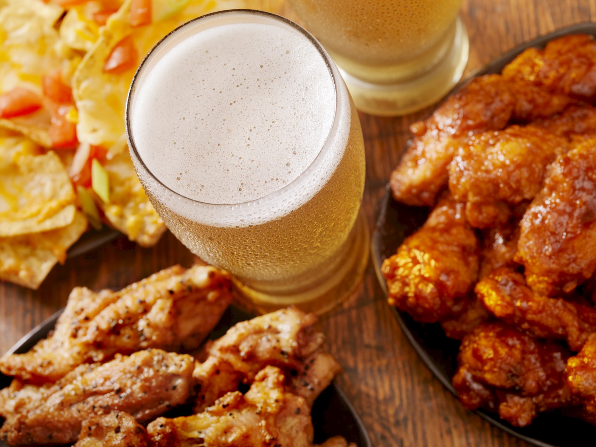 Nachos with toppings, two glasses of beer, and plates of chicken wings in different flavors on a wooden table.