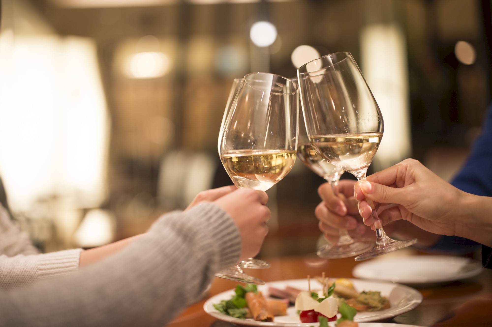 People clinking wine glasses over a table with appetizers, suggesting a celebratory or social gathering in a warm, dimly lit setting.