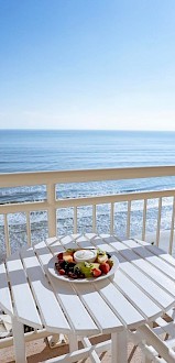 A white round table with fruit and yogurt on a balcony overlooking the beach and ocean under a bright, sunny sky.