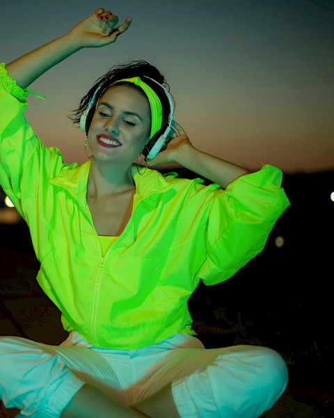 A person wearing a bright neon jacket and white pants sits outdoors at dusk, smiling with closed eyes and raised arms.