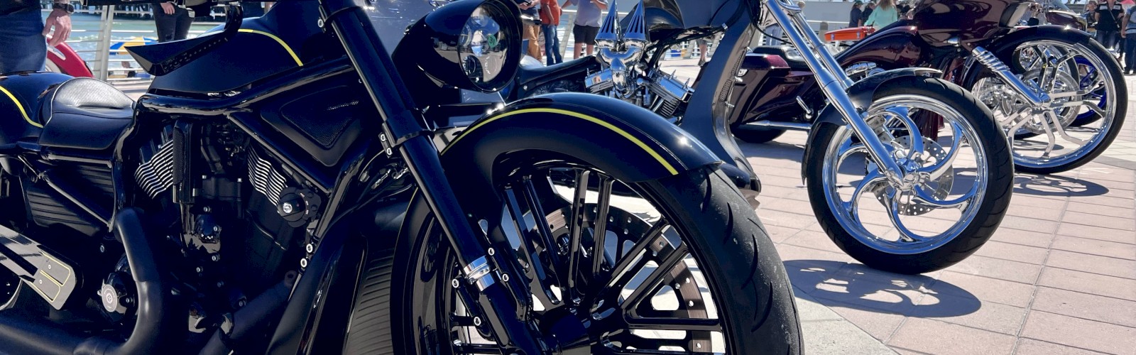 Custom motorcycles lined up outdoors with people in the background under a clear blue sky.