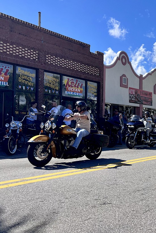 A street scene with many motorcyclists gathered in front of shops under a bright, partly cloudy sky in a tropical location.