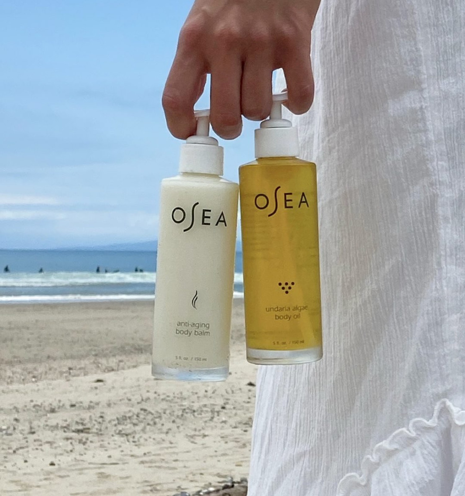 The image shows a person holding two bottles of beach skincare products, a body balm and body oil, with a beach and ocean in the background.