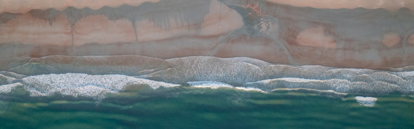 Aerial view of a beach with waves lapping a sandy shoreline and a few buildings visible at the top, meeting turquoise water below.