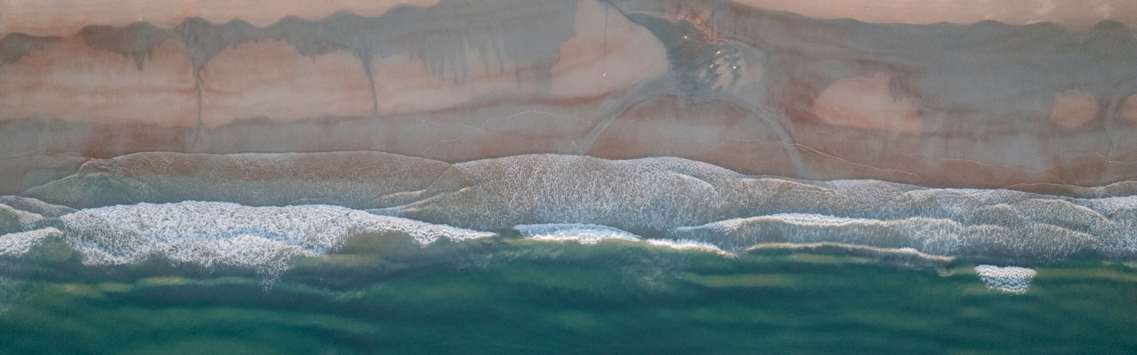 Aerial view of a beach with waves meeting the shore, and sand patterns visible.