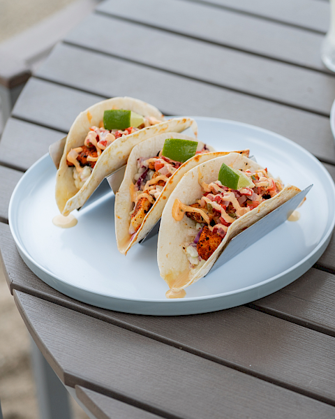 Three tacos filled with chicken, vegetables, and sauce, garnished with lime wedges, served on a white plate on a wooden table.