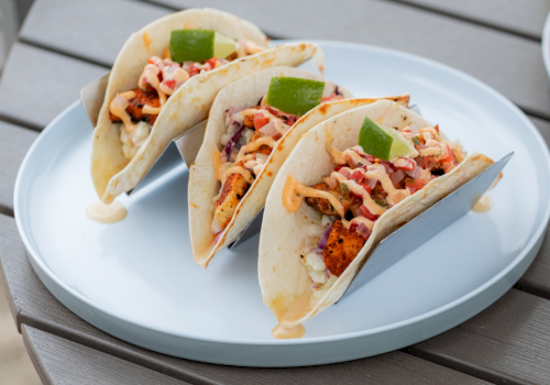 Three tacos filled with chicken, vegetables, and sauce, garnished with lime wedges, served on a white plate on a wooden table.