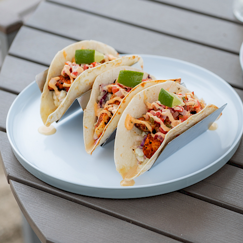 Three tacos filled with chicken, vegetables, and sauce, garnished with lime wedges, served on a white plate on a wooden table.