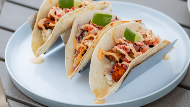 Three tacos filled with chicken, vegetables, and sauce, garnished with lime wedges, served on a white plate on a wooden table.