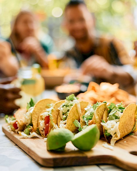 A group of people enjoy tacos on a wooden platter, with lime wedges and hot sauce bottles nearby on a sunny day.