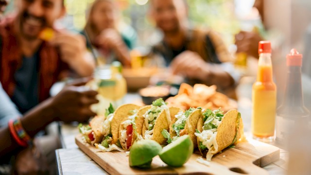 A group of people enjoy tacos on a wooden platter, with lime wedges and hot sauce bottles nearby on a sunny day.