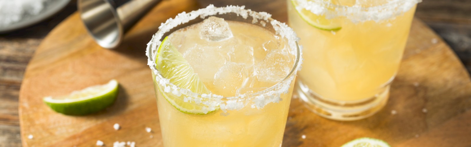 Two margarita glasses with lime slices and salted rims on a wooden board, accompanied by a metal jigger.
