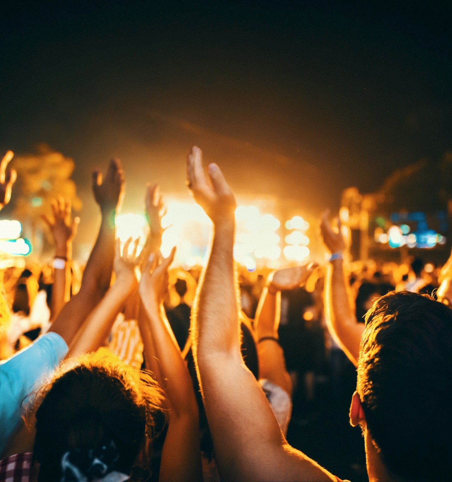 A crowd at a lively concert or festival, hands raised, surrounded by bright stage lights creating a vibrant and energetic atmosphere.