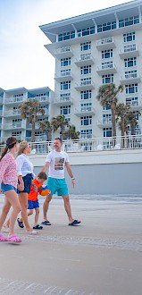 A family of four is walking on a beach promenade in front of a multi-story hotel under a partly cloudy sky.