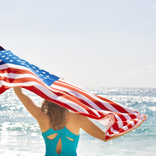 A person at the beach holds a U.S. flag in the sunlight, facing the ocean waves.