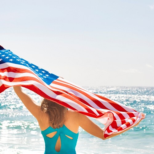 A woman holding an American flag on the beach during sunny weather with the ocean in the background.