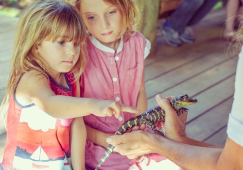 Two young girls observe and touch a lizard held by a person, engaging closely with nature on a wooden deck.