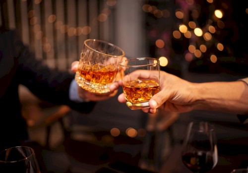 Two people clink glasses filled with amber liquid in a celebratory toast, with a festive bokeh background adding warmth to the scene.