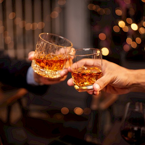 Two people clink glasses filled with amber liquid in a celebratory toast, with a festive bokeh background adding warmth to the scene.