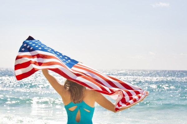 A person at the beach holds up the U.S. flag, wearing a turquoise swimsuit, facing the ocean under a sunny sky.
