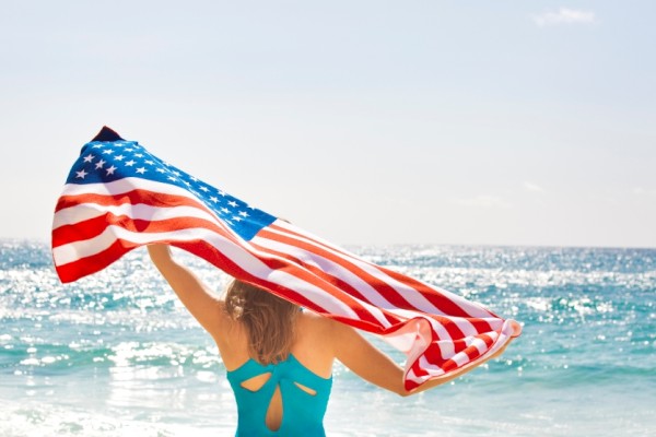 A woman holding an American flag on the beach with the ocean in the background, celebrating patriotism and summer.
