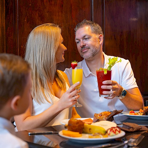 A couple is enjoying drinks at a table, with a child nearby. The table has breakfast items like muffins and a banana.