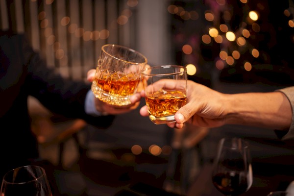 Two people clinking glasses of whiskey against a backdrop of warm, festive lights, creating a cozy and celebratory atmosphere.