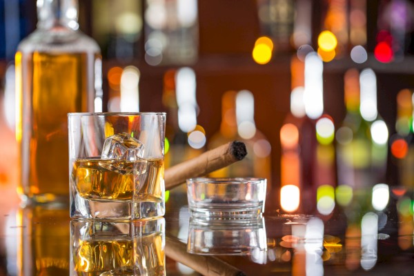 A glass of whiskey with ice, a cigar in an ashtray, and a bottle are placed on a bar counter with blurred lights in the background.