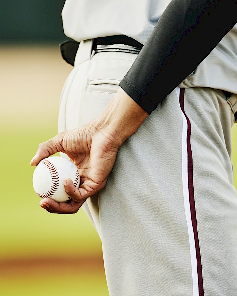 A person in a baseball uniform is holding a baseball behind their back on a field.