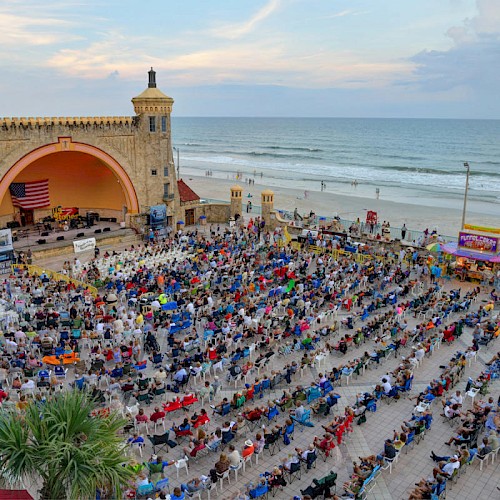 A large crowd is gathered by a seaside amphitheater with an arch, attending an event or concert, with the ocean visible in the background.