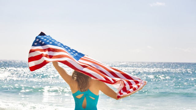 A person at the beach holds a U.S. flag in the sunlight, facing the ocean waves.